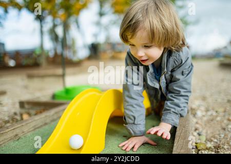 Mignon garçon en bas âge jouant au mini-golf s'amusant sur une aire de jeux en plein air le chaud jour d'automne. Loisirs actifs pour les enfants en automne. Banque D'Images