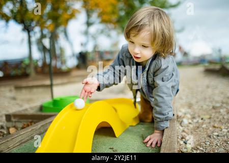 Mignon garçon en bas âge jouant au mini-golf s'amusant sur une aire de jeux en plein air le chaud jour d'automne. Loisirs actifs pour les enfants en automne. Banque D'Images