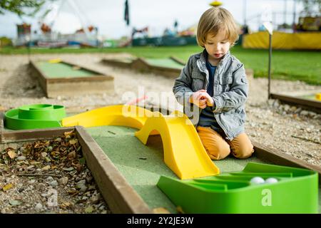 Mignon garçon en bas âge jouant au mini-golf s'amusant sur une aire de jeux en plein air le chaud jour d'automne. Loisirs actifs pour les enfants en automne. Banque D'Images