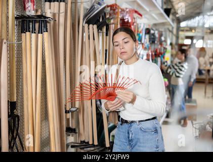 Jeune femme avec intérêt choisit le râteau dans la quincaillerie pour le jardinage Banque D'Images