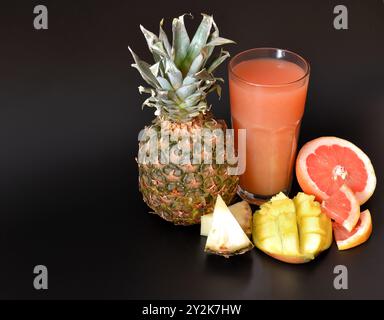 Un mélange de jus de fruits dans un grand verre à facettes sur fond noir, à côté de morceaux d'ananas mûrs, de pamplemousse et de mangue. Gros plan. Banque D'Images