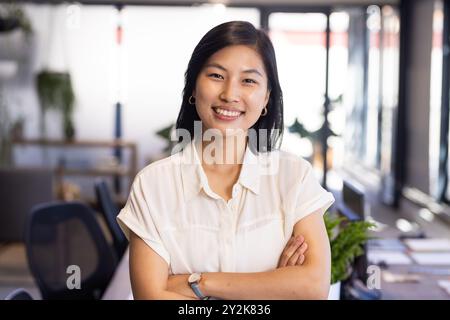 Debout dans un bureau moderne, femme asiatique confiante avec les bras croisés, souriant professionnellement Banque D'Images