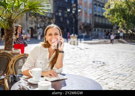 Une femme aux cheveux longs est assise à une table de café, souriant tout en parlant au téléphone. Elle aime une tasse de café et écrit dans un cahier, entourée de salut Banque D'Images