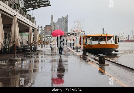 Hambourg, Allemagne. 11 septembre 2024. Un passant avec un parapluie marche le long du Landungsbrücken. La silhouette frappante de la salle de concert Elbphilharmonie est visible en arrière-plan. Crédit : Christian Charisius/dpa/Alamy Live News Banque D'Images