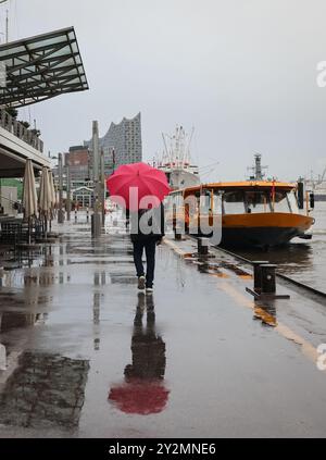 Hambourg, Allemagne. 11 septembre 2024. Un passant avec un parapluie marche le long du Landungsbrücken. La silhouette frappante de la salle de concert Elbphilharmonie est visible en arrière-plan. Crédit : Christian Charisius/dpa/Alamy Live News Banque D'Images