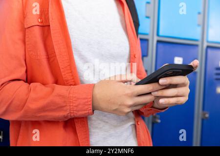 En utilisant un smartphone, adolescente debout près de casiers à l'école, portant une veste orange Banque D'Images