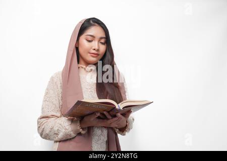 Femme musulmane asiatique sérieuse portant le voile hijab priant et lisant Al Coran, debout sur fond blanc isolé. Ramadan et Aïd Moubarak concept Banque D'Images