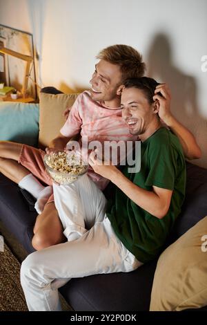 Deux hommes partagent rire et pop-corn tout en se relaxant ensemble à la maison. Banque D'Images