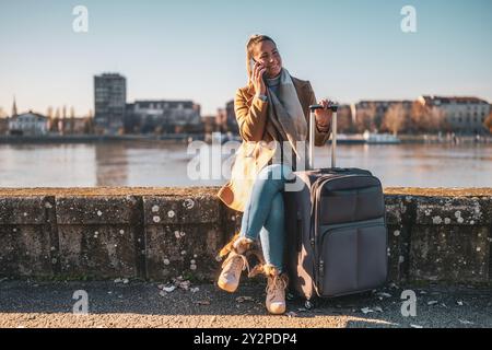 Femme heureuse touriste avec valise parlant au téléphone tout en se reposant au bord de la rivière. Image teintée. Banque D'Images