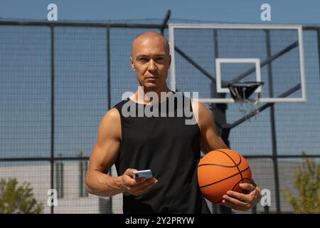 Sport homme caucasien ou joueur de basket à l'aide d'un smartphone après un entraînement en plein air. Collations musclées d'athlète sur une barre protéinée pendant le repos après l'entraînement Banque D'Images