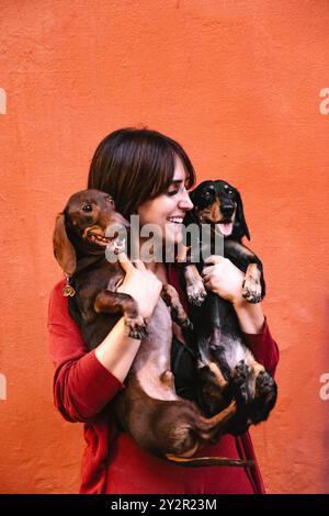 Une jeune femme joyeuse dans un pull rouge tient joyeusement deux chiens Teckel sur un fond orange vif les chiens, à l'air ludique, ajoutent à la chaleur Banque D'Images