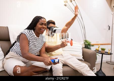 Femme noire joyeuse et son partenaire ayant une session de jeu amusante dans leur salon confortable, elle rit comme il joue avec un casque VR Banque D'Images