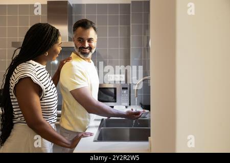 Un couple joyeux se tient près de l'évier de la cuisine, partageant un moment de lumière la femme, une femme noire, sourit à son partenaire, un indien avec une barbe Banque D'Images