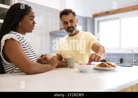 Une femme noire et un homme indien assis à leur comptoir de cuisine lumineux, profitant d'un petit déjeuner confortable ensemble, ils discutent autour de tasses de café et de croissants, Banque D'Images