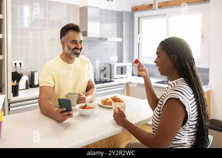 Un couple joyeux s'engage dans une conversation matinale animée, partageant des sourires et un petit déjeuner sain dans une cuisine moderne et lumineuse Banque D'Images