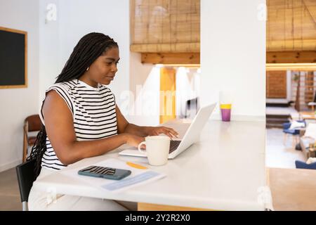 Femme noire travaillant confortablement sur un ordinateur portable dans une maison ensoleillée et moderne, reflétant un équilibre harmonieux travail-vie privée Banque D'Images