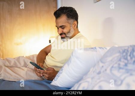 Souriant homme indien confortablement en utilisant son téléphone tout en étant allongé dans le lit, démontrant la technologie dans la vie domestique quotidienne Banque D'Images