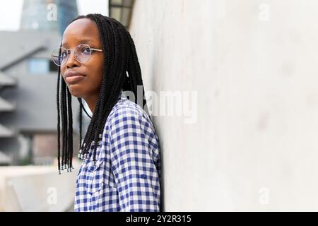 Une femme noire se tient réfléchie dans un quartier d'affaires, son regard introspectif portant des lunettes et une chemise à carreaux, elle reflète une moderne, la professionnona Banque D'Images