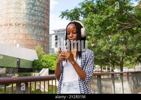 Une jeune femme noire aime écouter de la musique à travers ses écouteurs tout en regardant son smartphone dans un quartier d'affaires animé, entouré d'architectu moderne Banque D'Images