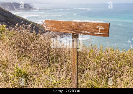 Un panneau rustique en bois affichant les directions de randonnée dans les Asturies, en Espagne, se dresse sur un fond de la côte pittoresque et de la mer turquoise. Banque D'Images