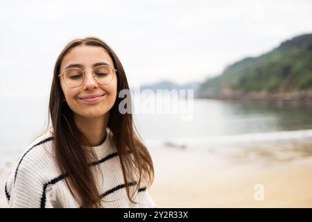 Jeune femme aux yeux fermés, souriante avec satisfaction, se tient au bord de la mer le long de la côte des Asturies, en Espagne, dégageant une aura paisible et réfléchissante au milieu d'un Banque D'Images