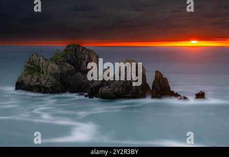 Un coucher de soleil vibrant illumine le ciel au-dessus des falaises rocheuses et des eaux de mer douces et tourbillonnantes au large de la côte de Liencres, Cantabrie en Espagne. Banque D'Images