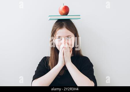 Adolescente avec une expression ludique équilibre un livre et une pomme sur sa tête tout en tenant ses mains ensemble devant son visage. Elle regarde Banque D'Images