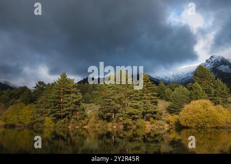 Un lac serein reflète les couleurs vives des arbres d'automne et une montagne enneigée sous un ciel orageux, créant un contraste et une profondeur étonnants dans le L. Banque D'Images