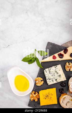 Vue de dessus d'un plateau de fromage élégant avec des variétés de fromage, noix, fruits secs, huile d'olive et basilic frais sur un fond blanc marbré, ide Banque D'Images