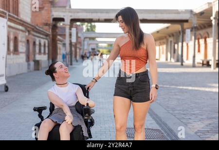 Deux jeunes femmes profitant d’une journée de sortie, avec une amie en fauteuil roulant, toutes deux partageant un rire dans un cadre urbain ensoleillé Banque D'Images