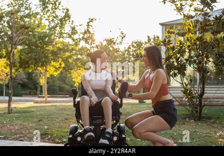 Un moment joyeux capturé alors que deux jeunes femmes partagent des rires dans un parc, dont l’une utilise un fauteuil roulant, soulignant l’inclusivité et l’amitié Banque D'Images