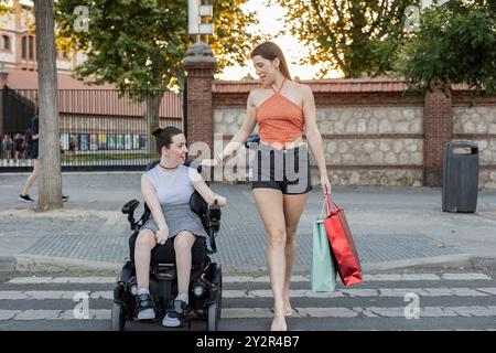 Deux jeunes femmes aiment faire du shopping dans la ville, dont l'une utilise un fauteuil roulant motorisé, elles parlent et sourient, mettant en valeur un positif et inclus Banque D'Images