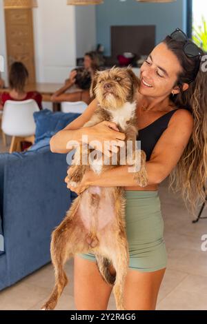 Une femme sourit en tenant un petit chien, le regardant affectueusement. En arrière-plan, d'autres participants sont engagés dans des activités intérieures. Banque D'Images