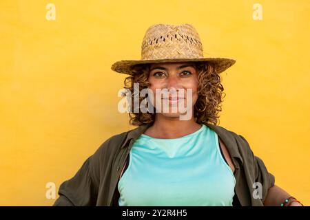 Une femme aux cheveux bouclés et un chapeau de paille sourit doucement à la caméra. Le fond est jaune vif, mettant en évidence l'expression joyeuse et l'été v Banque D'Images