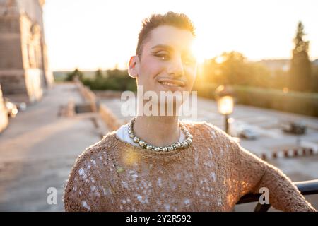 Une personne rayonnante non binaire partage un sourire confiant à la caméra, se prélassant dans le soleil chaud de l'heure dorée avec un arrière-plan urbain flou. Banque D'Images