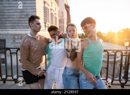 Un groupe d'amis LGBTQIA+ divers partagent des rires embrasser et danser devant un coucher de soleil urbain, rayonnant de positivité et de compagnie. Banque D'Images