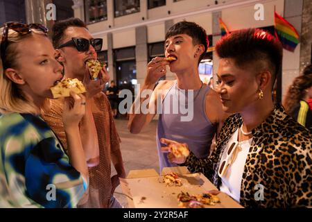 Un groupe diversifié d'amis LGBTQIA+ partagent une pizza dans une rue de la ville la nuit, célébrant l'amitié et l'identité. Banque D'Images