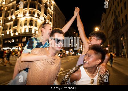 Un groupe joyeux d’amis LGBTQIA+ profitent d’une soirée animée en ville, riant et se soulevant. L'arrière-plan urbain bien éclairé offre un Banque D'Images