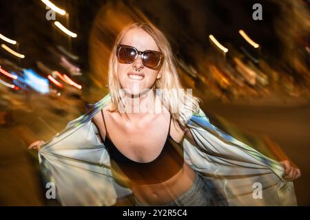 Une jeune femme élégante prend la pose dans une rue de la ville la nuit, avec des lumières floues créant un arrière-plan dynamique Banque D'Images