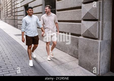 Un couple gay multiethnique marche main dans la main, partageant un rire en se promenant dans une rue de la ville, exsudant confort et bonheur en compagnie de l'autre. Banque D'Images