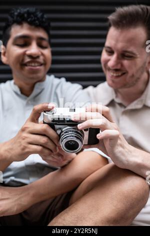 Un joyeux couple gay multiethnique partage un rire tout en examinant des photos sur un appareil photo vintage, assis étroitement ensemble dans un cadre décontracté. Banque D'Images