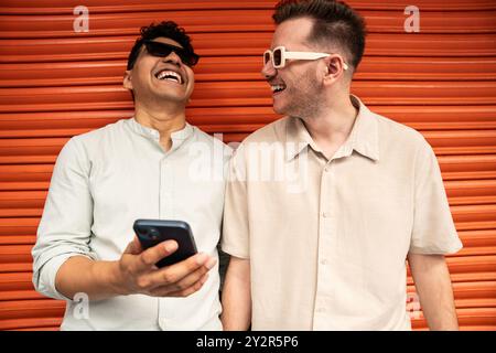 Un joyeux couple gay multiethnique partage un moment de lumière sur un fond orange vif. Un homme tient un smartphone, partageant peut-être un somethi Banque D'Images