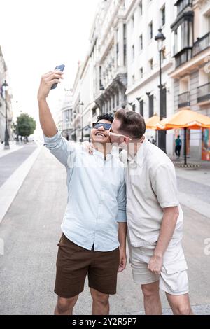 Un joyeux couple gay multiethnique partage un moment ludique tout en prenant un selfie dans une rue animée de la ville, avec un partenaire embrassant la joue de l'autre. Banque D'Images