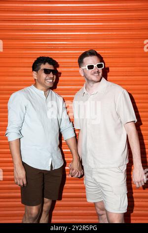 Un couple gay multiethnique affectueux et joyeux se tenant la main, souriant largement devant un fond orange vibrant. Les deux portent des lunettes de soleil A. Banque D'Images