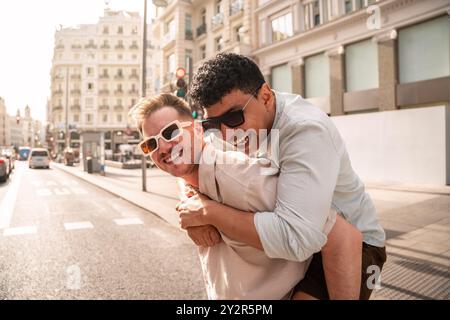 Un joyeux couple gay multiethnique partage un tour de piggyback, riant ensemble dans la lumière du soleil éclatante d'une rue animée de la ville, rayonnant d'amour et de bonheur Banque D'Images