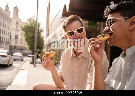 Un joyeux couple gay multiethnique partage un moment de lumière en mangeant des beignets dans une rue urbaine ensoleillée, reflétant le bonheur et la vie urbaine détendue. Banque D'Images