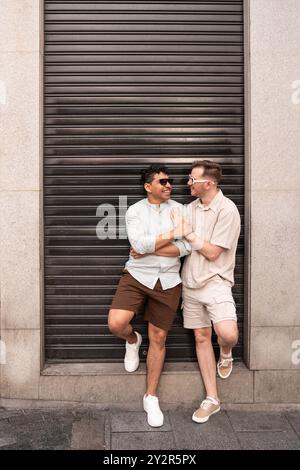 Un joyeux couple gay multiethnique partage un rire tout en s’engageant ludique les uns avec les autres devant un volet fermé, montrant de l’affection et du bonheur. Banque D'Images