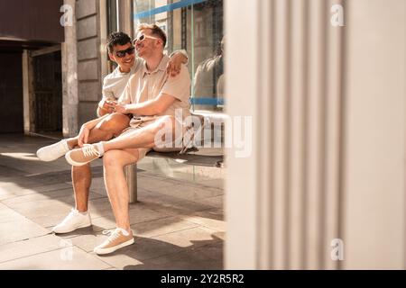 Un joyeux couple gay multiethnique se détend sur un banc d'arrêt de bus sous la lumière chaude du soleil, souriant et s'embrassant dans un moment franc et affectueux Banque D'Images