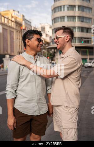 Un couple gay multiethnique partage un moment joyeux dans une rue de la ville, souriant et s'embrassant chaleureusement dans une tenue d'été décontractée. Banque D'Images