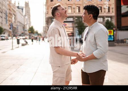 Un couple gay multiethnique partage un moment joyeux, se tenant la main et se souriant dans une rue de la ville, dépeignant l'amour et la connexion dans une ville Banque D'Images